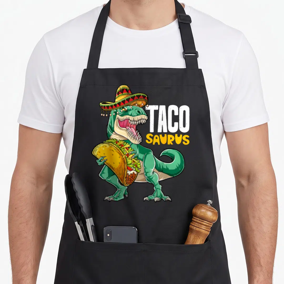 Taco dinosaur apron - Green T-Rex in sombrero holding taco with TACO SAURUS text — shown on a black cotton canvas apron against a white background