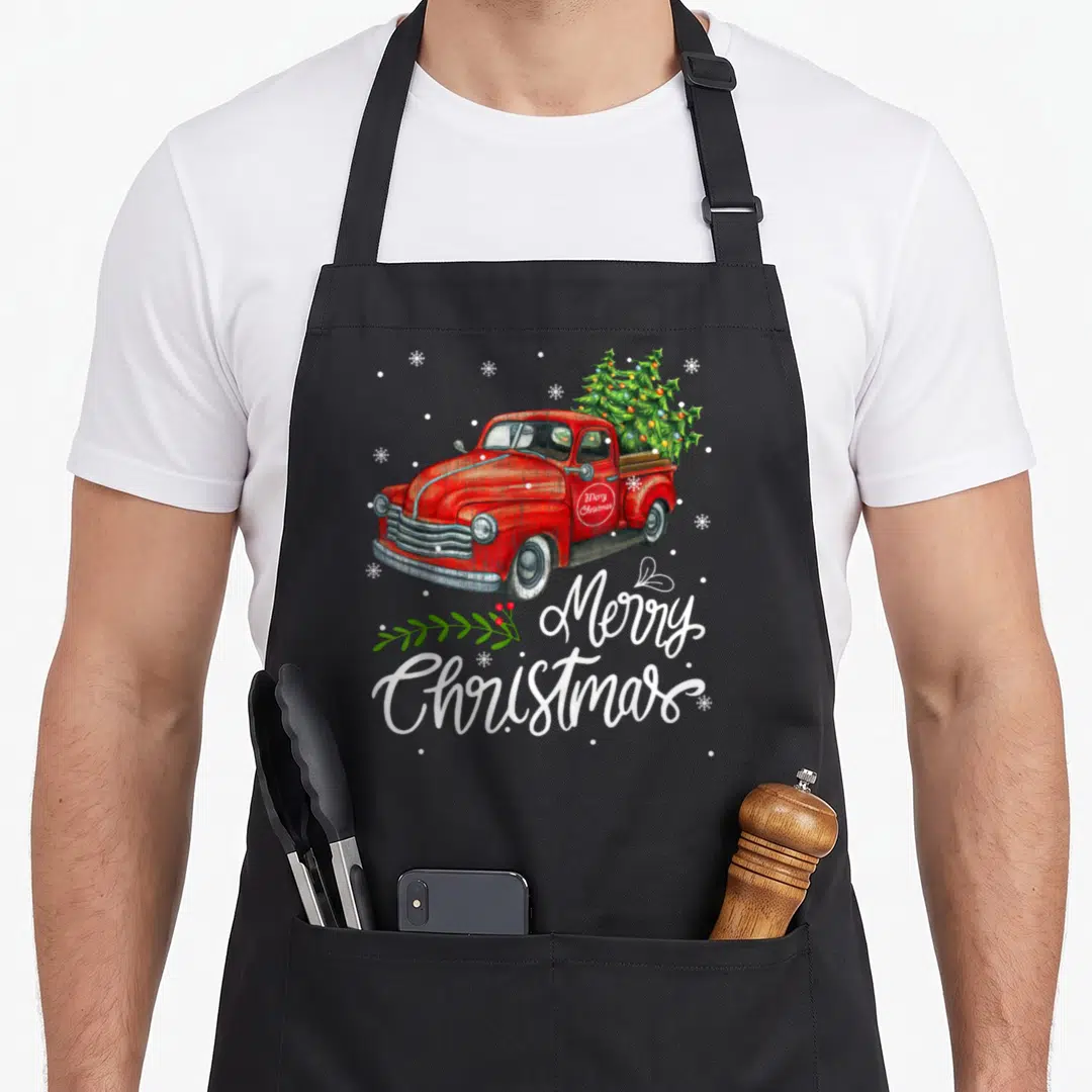 Pajama christmas tree apron - Red truck carrying three decorated Christmas trees with white Merry Christmas script — shown on a black cotton canvas apron against a white background