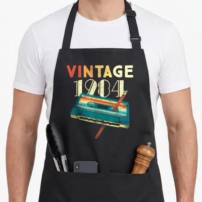 Cassette 42nd birthday apron - Retro cassette tape with pencil and stacked vintage 1984 text in teal orange cream — shown on a black cotton canvas apron against a white background