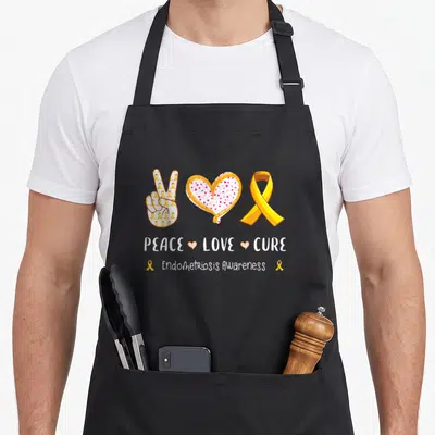 Peace endometriosis awareness apron - Cartoon peace hand pink heart yellow ribbon PEACE LOVE CURE Endometriosis — shown on a black cotton canvas apron against a white background