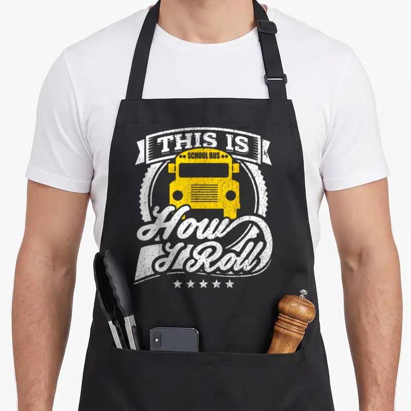 Roll school bus apron - Yellow school bus with This Is How I Roll distressed text and five stars — shown on a black cotton canvas apron against a white background