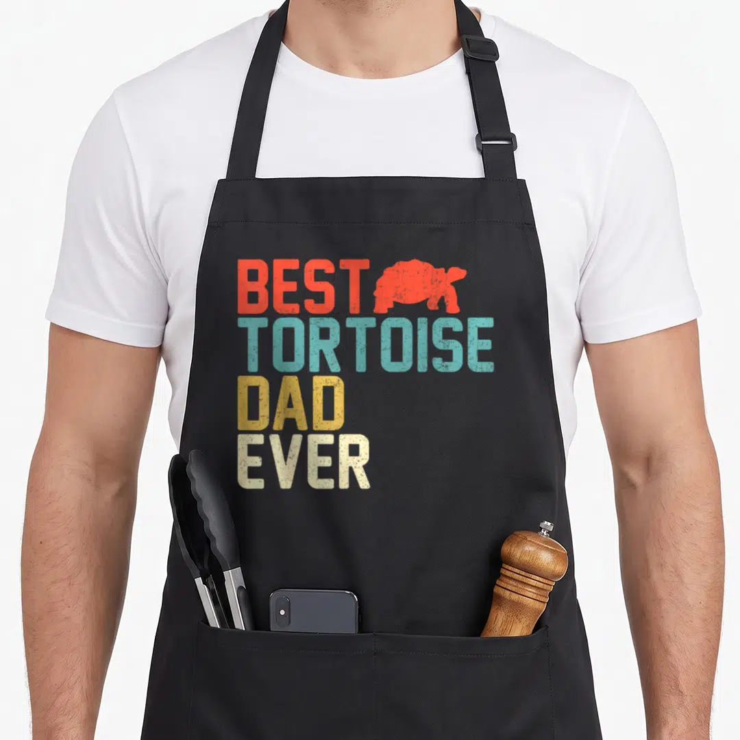 Tortoise turtle dad apron - Red tortoise silhouette beside stacked BEST TORTOISE DAD EVER text in teal yellow and beige — shown on a black cotton canvas apron against a white background