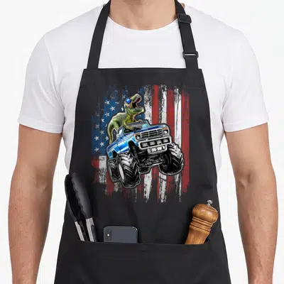 Dinosaur 4th of july apron - Green T-Rex in blue monster truck with U.S.A. plate on distressed flag — shown on a black cotton canvas apron against a white background
