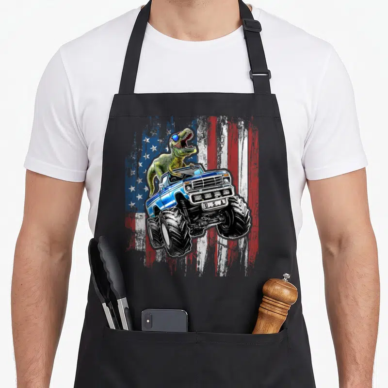Dinosaur 4th of july apron - Green T-Rex in blue monster truck with U.S.A. plate on distressed flag — shown on a black cotton canvas apron against a white background