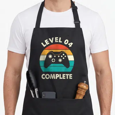 Level complete wedding aprons - Retro game controller inside striped sun with LEVEL 04 COMPLETE text — shown on a black cotton canvas apron against a white background