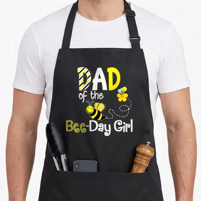Hive bee apron - DAD of the Bee-Day Girl text with two cartoon bees and dotted flight path — shown on a black cotton canvas apron against a white background