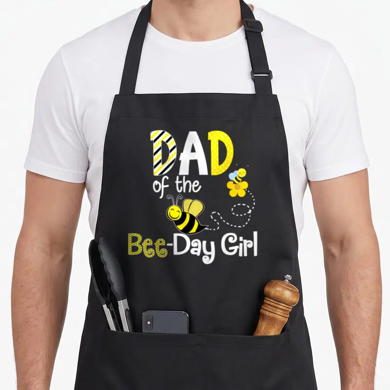 Hive bee apron - DAD of the Bee-Day Girl text with two cartoon bees and dotted flight path — shown on a black cotton canvas apron against a white background