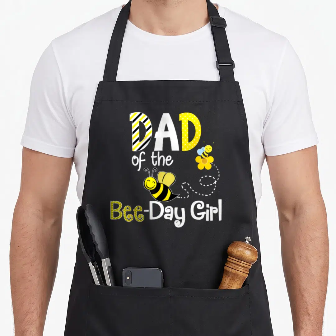 Hive bee apron - DAD of the Bee-Day Girl text with two cartoon bees and dotted flight path — shown on a black cotton canvas apron against a white background