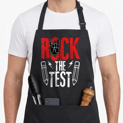 Metal teacher apron - Red ROCK text with black hand symbol, white THE TEST flanked by lightning bolts and pencils — shown on a black cotton canvas apron against a white background