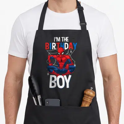 Funny animal birthday apron - Spider-Man crouching under I'M THE BIRTHDAY BOY text with white web and small spiders — shown on a black cotton canvas apron against a white background