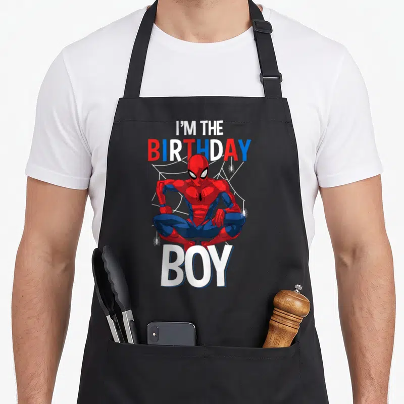 Funny animal birthday apron - Spider-Man crouching under I'M THE BIRTHDAY BOY text with white web and small spiders — shown on a black cotton canvas apron against a white background