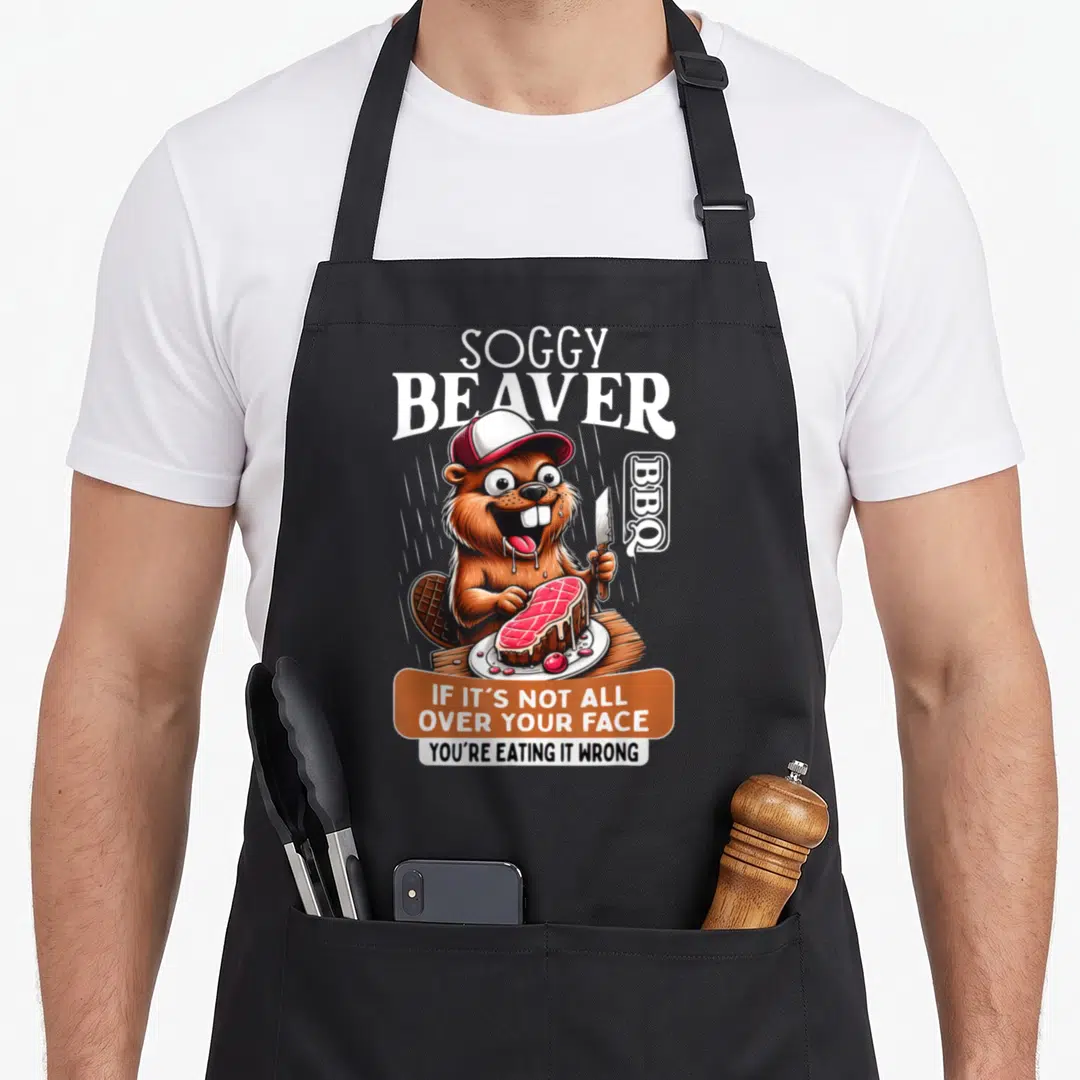Barbecue beaver apron - Cartoon brown beaver wearing cap eating red steak with white and red text — shown on a black cotton canvas apron against a white background
