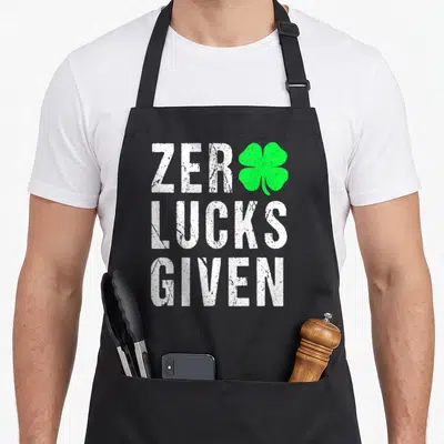 Saint patrick irish aprons - White distressed text reading Zero Lucks Given with a green four-leaf clover replacing — shown on a black cotton canvas apron against a white background
