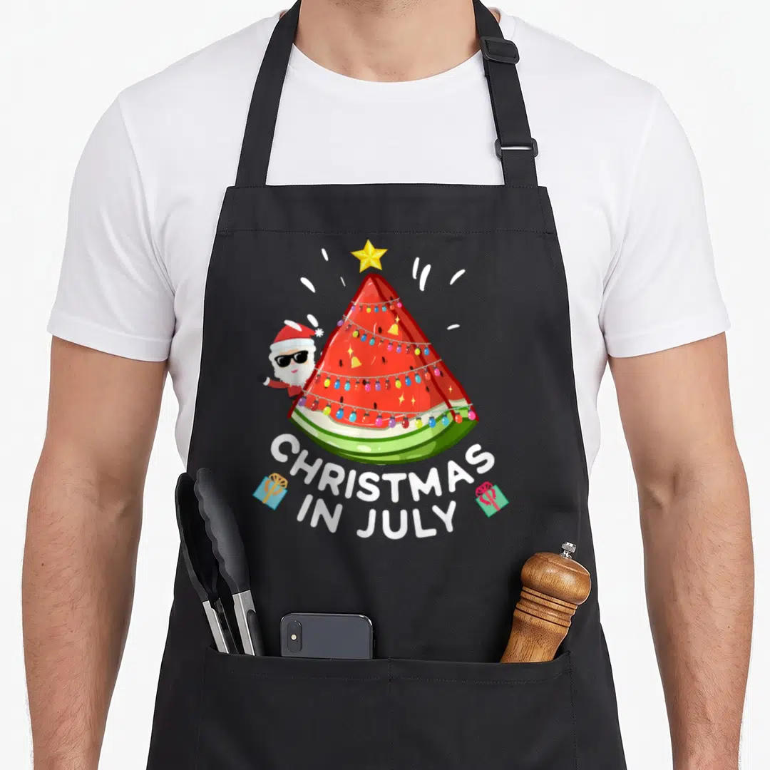 July watermelon apron - Watermelon tree with Santa, lights, and CHRISTMAS IN JULY text flanked by gift boxes — shown on a black cotton canvas apron against a white background