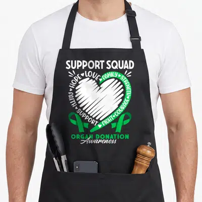Support organ apron - White heart surrounded by support words and two green ribbons for organ donation — shown on a black cotton canvas apron against a white background