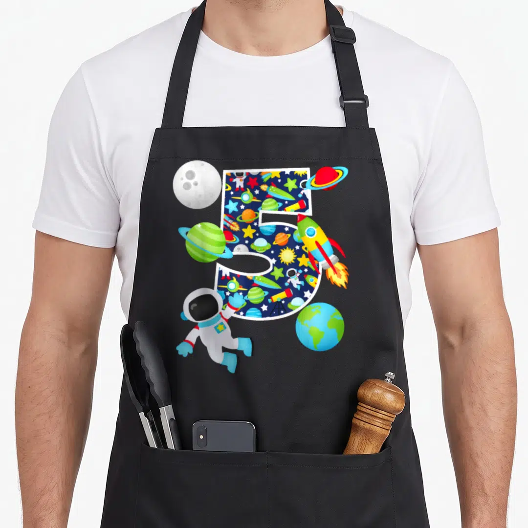 System birthday apron - Number 5 filled with space pattern surrounded by astronaut rocket and planets in cartoon style — shown on a black cotton canvas apron against a white background