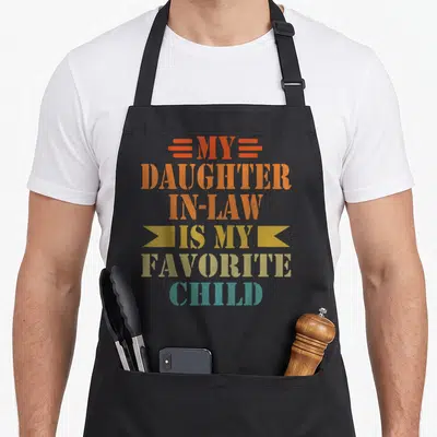 Cute law father day apron - Stacked orange yellow teal text MY DAUGHTER IN-LAW IS MY FAVORITE CHILD with decorative bars — shown on a black cotton canvas apron against a white background