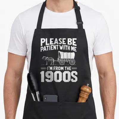 Patient 1900s apron - White covered wagon with driver and PLEASE BE PATIENT WITH ME I'M FROM THE 1900S text — shown on a black cotton canvas apron against a white background