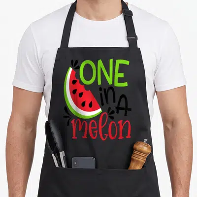 Adorable watermelon apron - Cartoon watermelon slice with ONE in A melon text in green, red, and black — shown on a black cotton canvas apron against a white background