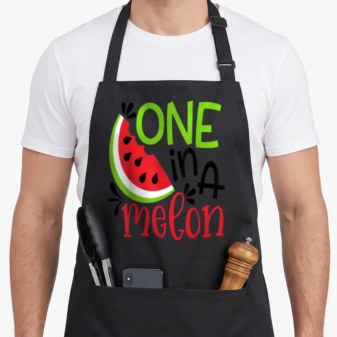 Adorable watermelon apron - Cartoon watermelon slice with ONE in A melon text in green, red, and black — shown on a black cotton canvas apron against a white background