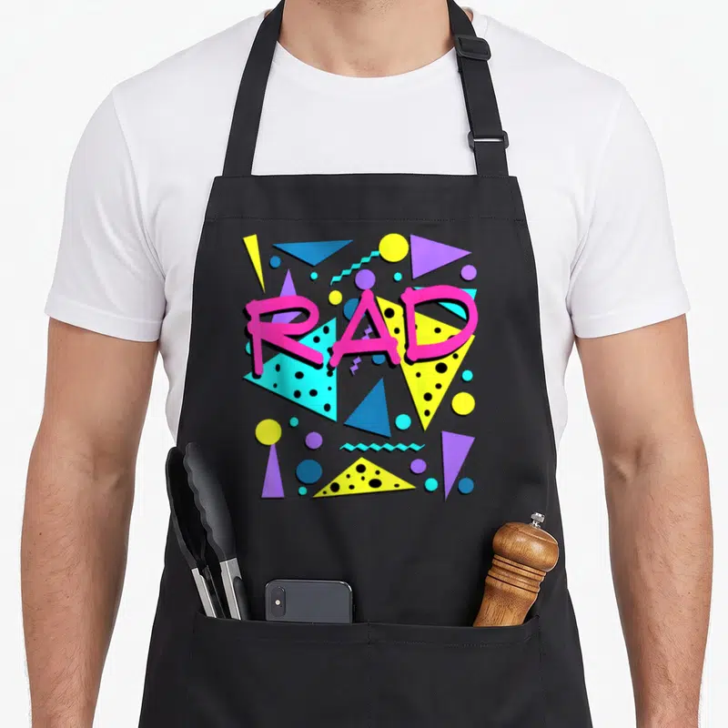 Rad 1980s apron - Pink RAD text over yellow blue and purple geometric triangles and circles — shown on a black cotton canvas apron against a white background