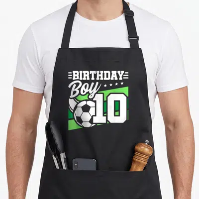 Soccer 10th birthday apron - White BIRTHDAY Boy 10 text and soccer ball on green geometric background — shown on a black cotton canvas apron against a white background