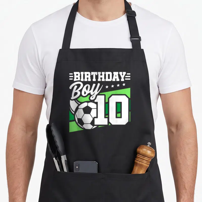 Soccer 10th birthday apron - White BIRTHDAY Boy 10 text and soccer ball on green geometric background — shown on a black cotton canvas apron against a white background
