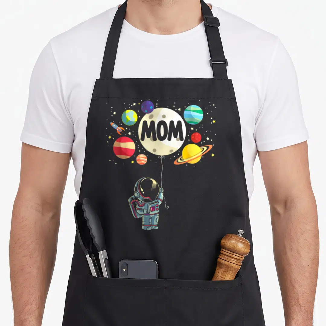 Astronaut family aprons - Cartoon astronaut holds MOM moon balloon among planets and stars on grey background — shown on a black cotton canvas apron against a white background
