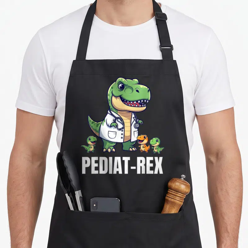 Pediatric t-rex apron - Green T-Rex in lab coat with two small dinos and PEDIAT-REX text — shown on a black cotton canvas apron against a white background