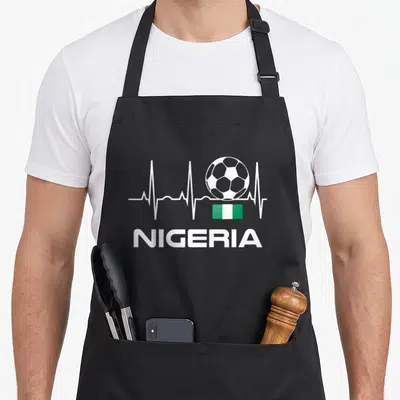 Nigeria football apron - Soccer ball on white heartbeat line above green and white Nigerian flag with NIGERIA text below — shown on a black cotton canvas apron against a white background