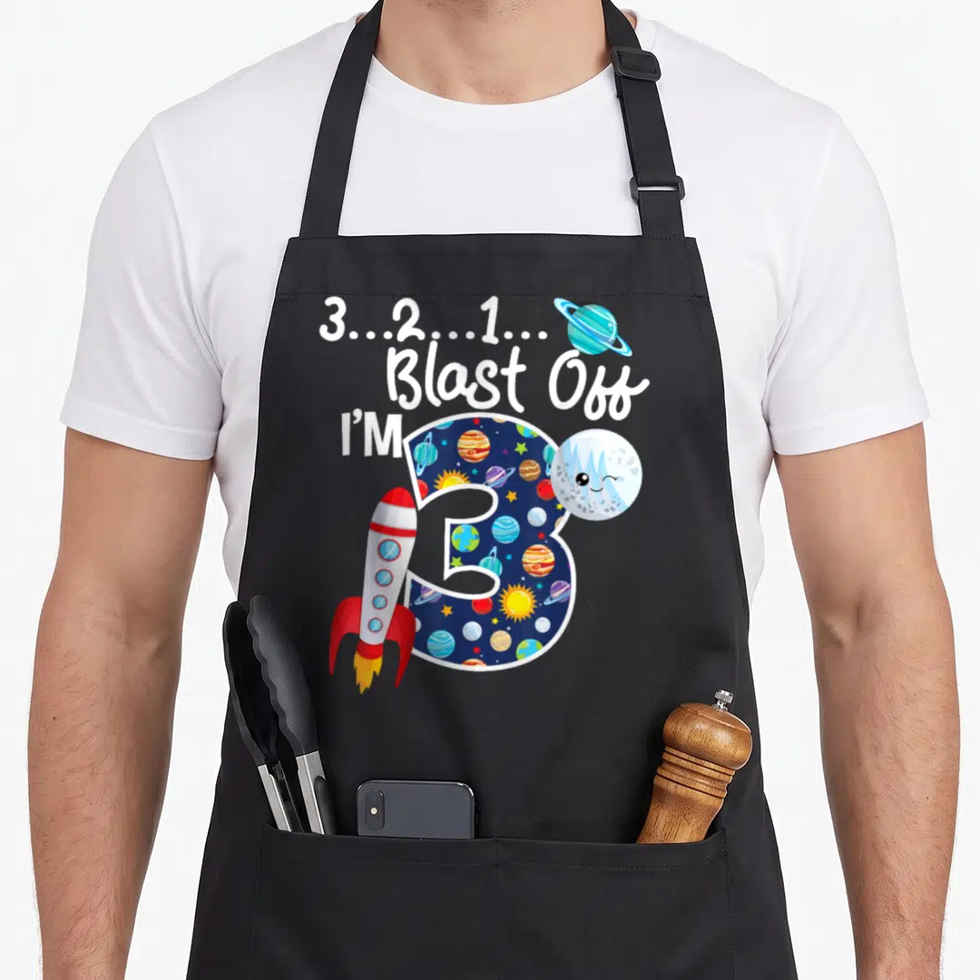 Outer space birthday apron - Number 3 filled with planets, rocket, winking planet, and blast off text in blue white red — shown on a black cotton canvas apron against a white background