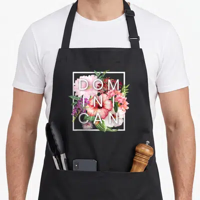 Flower dominican pride apron - White DOMINICAN text over pink and red watercolor flowers in a white square frame — shown on a black cotton canvas apron against a white background