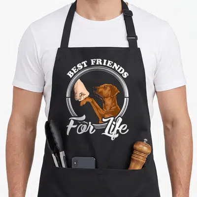 Retro rhodesian ridgeback apron - Cartoon brown dog shaking human paw inside white circle with BEST FRIENDS For Life — shown on a black cotton canvas apron against a white background