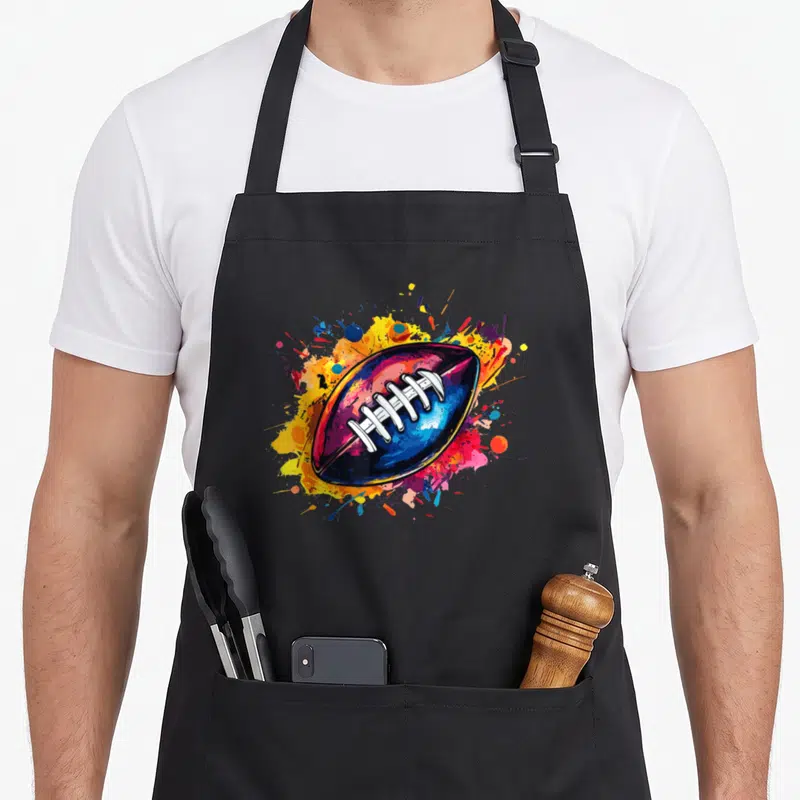 Game player football apron - Colorful cartoon American football on blue, yellow, red, and purple paint splatters — shown on a black cotton canvas apron against a white background