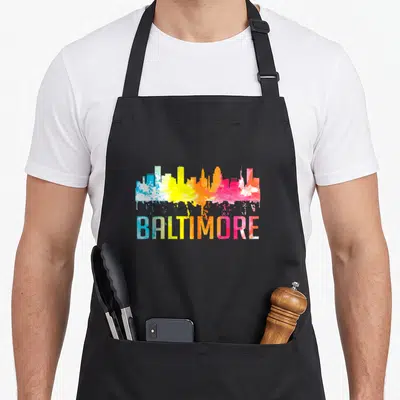 Baltimore skyline art apron - Watercolor Baltimore skyline in blue yellow orange pink with paint drips below text — shown on a black cotton canvas apron against a white background