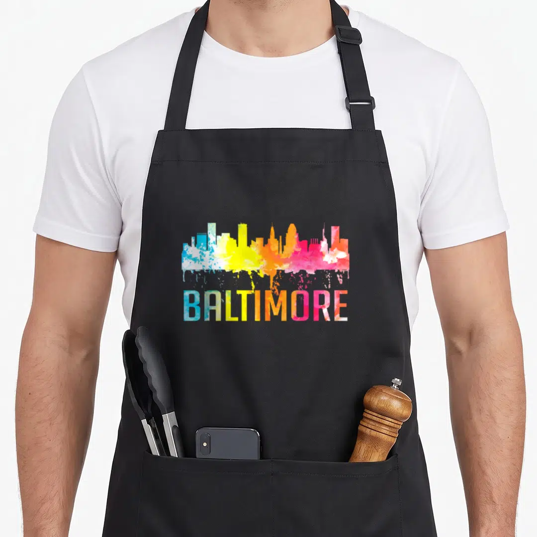Baltimore skyline art apron - Watercolor Baltimore skyline in blue yellow orange pink with paint drips below text — shown on a black cotton canvas apron against a white background
