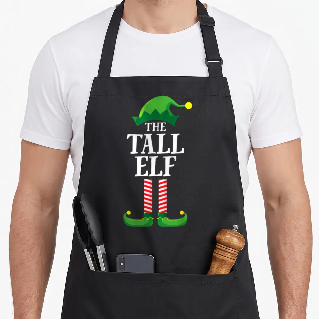 Tall christmas elf apron - Cartoon elf hat and striped legs framing stacked white text reading THE TALL ELF — shown on a black cotton canvas apron against a white background