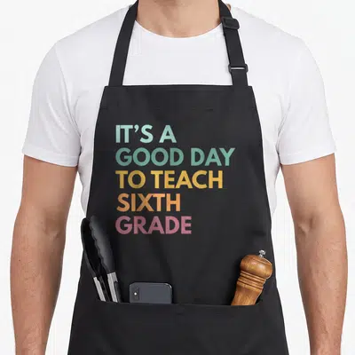 Sixth art teacher aprons - Stacked colorful text reading IT'S A GOOD DAY TO TEACH SIXTH GRADE in teal yellow orange pink — shown on a black cotton canvas apron against a white background