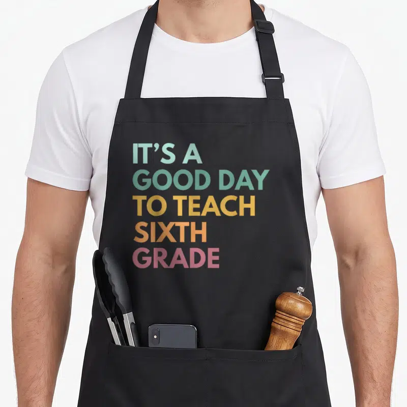 Sixth art teacher aprons - Stacked colorful text reading IT'S A GOOD DAY TO TEACH SIXTH GRADE in teal yellow orange pink — shown on a black cotton canvas apron against a white background
