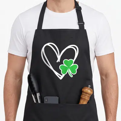 Toddler irish aprons - White outlined heart with green shamrock overlapping bottom right corner — shown on a black cotton canvas apron against a white background