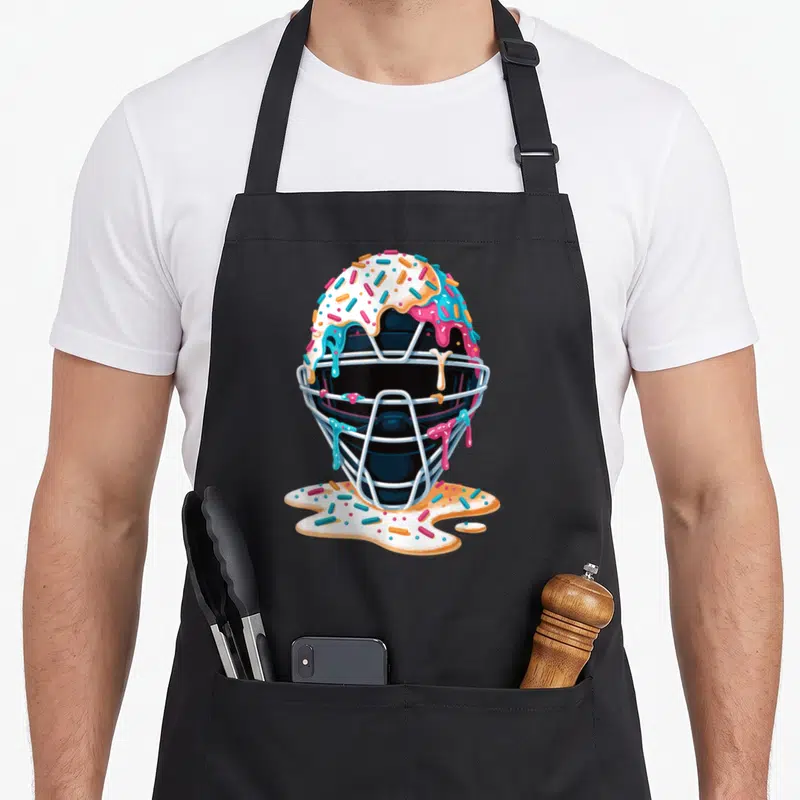 Catcher ice cream apron - Black catcher mask covered in dripping white pink blue icing and multicolored sprinkles — shown on a black cotton canvas apron against a white background