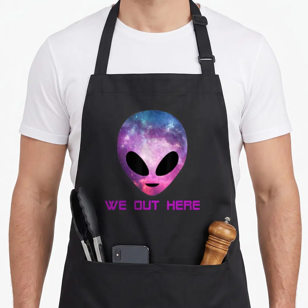 Trippy world music apron - Galaxy alien head in purple and blue above pink WE OUT HERE text — shown on a black cotton canvas apron against a white background