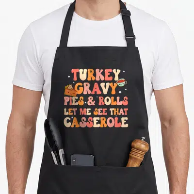 See thanksgiving apron - Retro cartoon text TURKEY GRAVY PIES ROLLS LET ME SEE THAT CASSEROLE with smiling pie — shown on a black cotton canvas apron against a white background