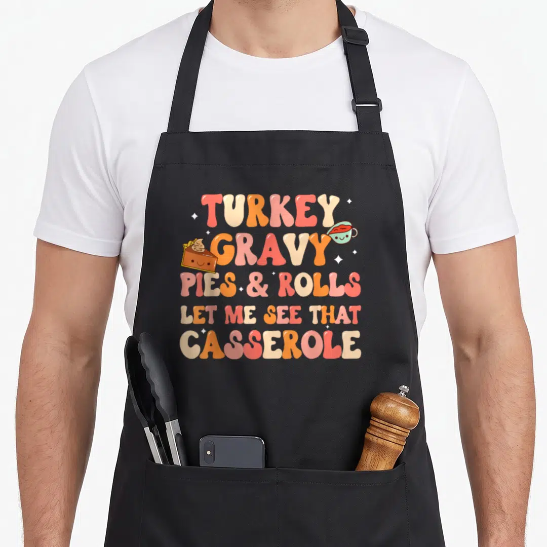 See thanksgiving apron - Retro cartoon text TURKEY GRAVY PIES ROLLS LET ME SEE THAT CASSEROLE with smiling pie — shown on a black cotton canvas apron against a white background