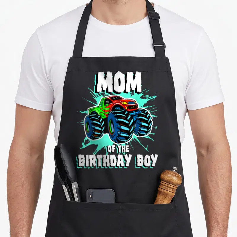 Funny birthday monster truck apron - MOM OF THE BIRTHDAY BOY text over red monster truck with blue tires — shown on a black cotton canvas apron against a white background