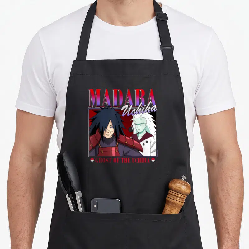 Shippuden 90s apron - MADARA Uchiha text with two anime characters and Ghost of the Uchiha flags — shown on a black cotton canvas apron against a white background