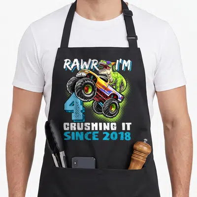 Dinosaur 4th truck apron - Green dinosaur driving monster truck over blue number 4 with arched text RAWR I'M — shown on a black cotton canvas apron against a white background