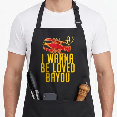 Loved crawfish boil apron - Red cartoon lobster above yellow I WANNA BE LOVED BAYOU stacked text — shown on a black cotton canvas apron against a white background