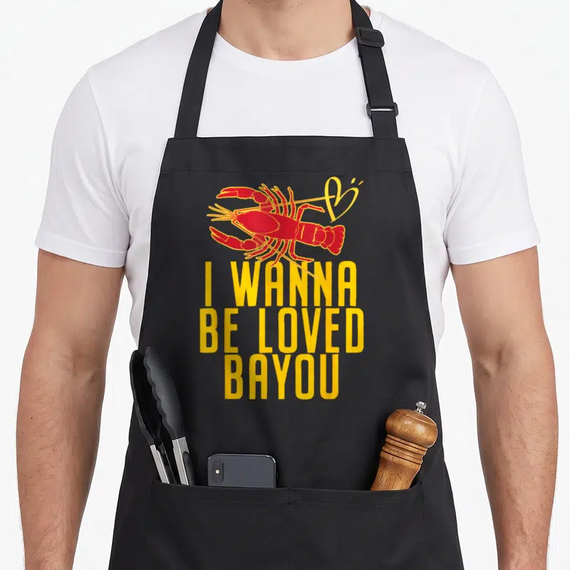 Loved crawfish boil apron - Red cartoon lobster above yellow I WANNA BE LOVED BAYOU stacked text — shown on a black cotton canvas apron against a white background