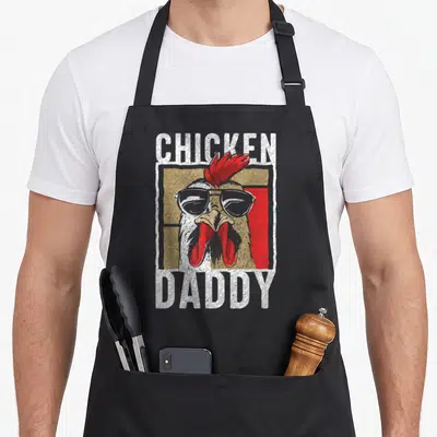 Daddy rooster apron - Cartoon rooster head with sunglasses and CHICKEN DADDY text in white red tan grey — shown on a black cotton canvas apron against a white background